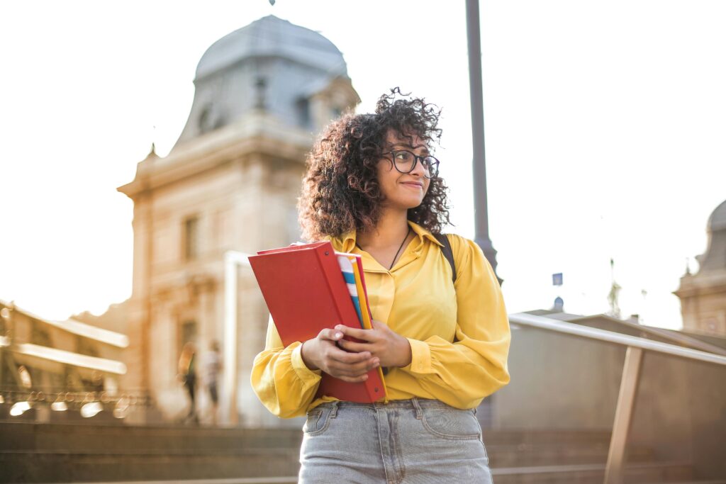 pexels-photo-3762800-3762800 Smiling female student with curly hair and glasses holding books on campus steps.